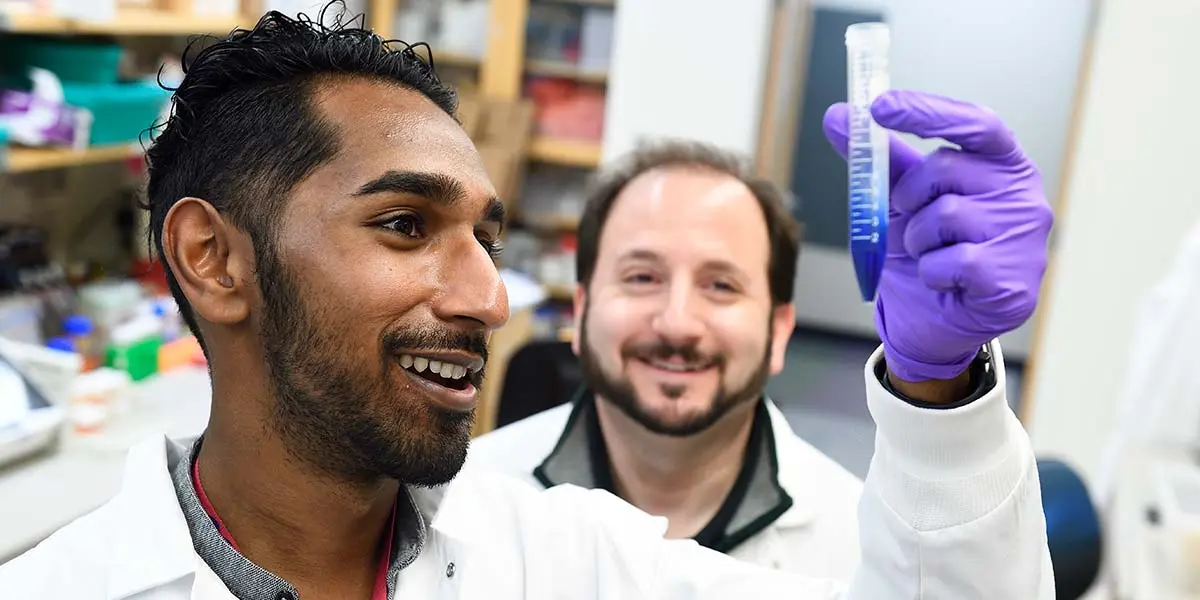 Two smiling scientists in white lab coats examine a blue liquid sample in a conical tube