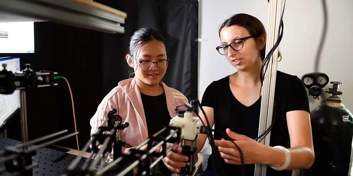 Two women work together on an optical laboratory setup with lasers, wires, and instruments in a research lab